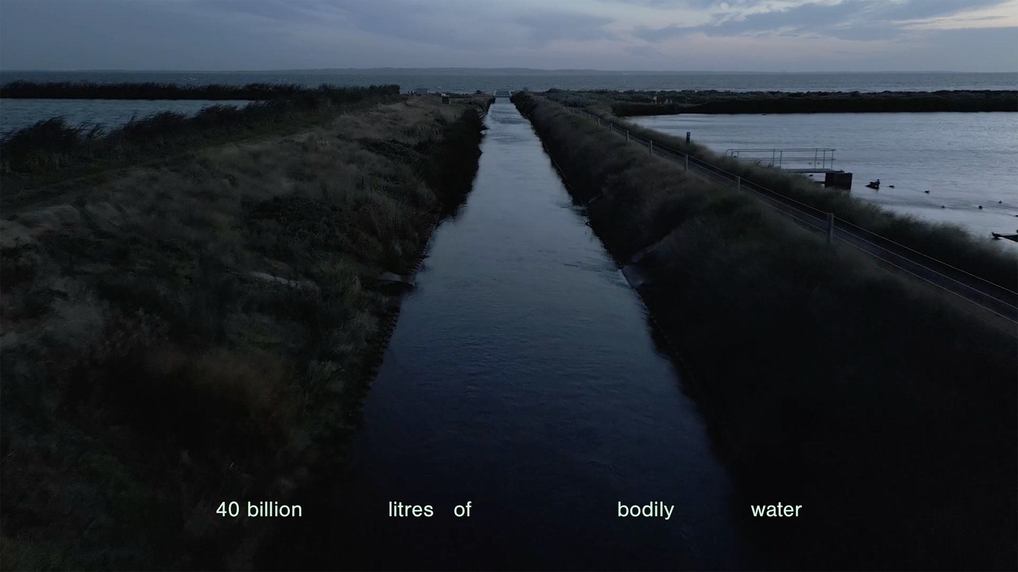 Video still from 'Metabolism' showing the 'open carrier', a channel of water, running out to the Port Phillip Bay at dusk. Text at the base of the frame says '40 billion litres of bodily water'.