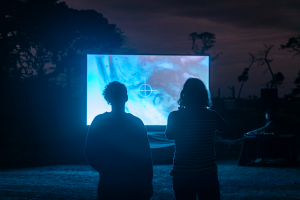 Two people stand in front of a bright screen at dusk, their backs to the camera. This is documentation of the screening of a work-in-progress version of Metabolism for 'Treatment III', Western Treatment Plant, Wadawurrung country, 2023.